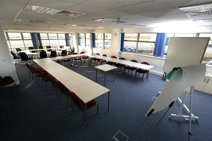 Empty training room set up with rows of tables and red chairs, a whiteboard easel in the foreground, and large windows