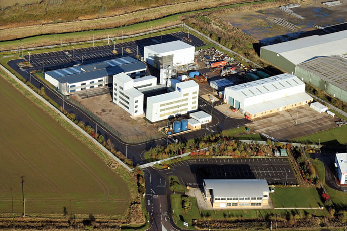 Aerial view of an industrial training site with white and grey buildings, parking areas, and green fields around it
