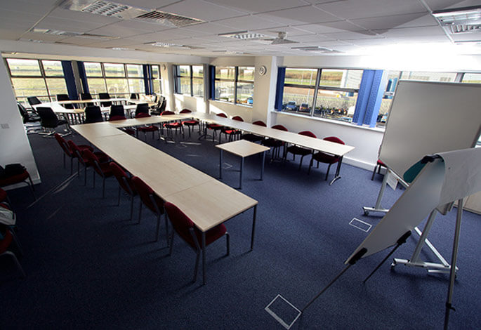 Empty conference room with tables arranged in a U-shape, red chairs, a whiteboard, and large windows with blue curtains